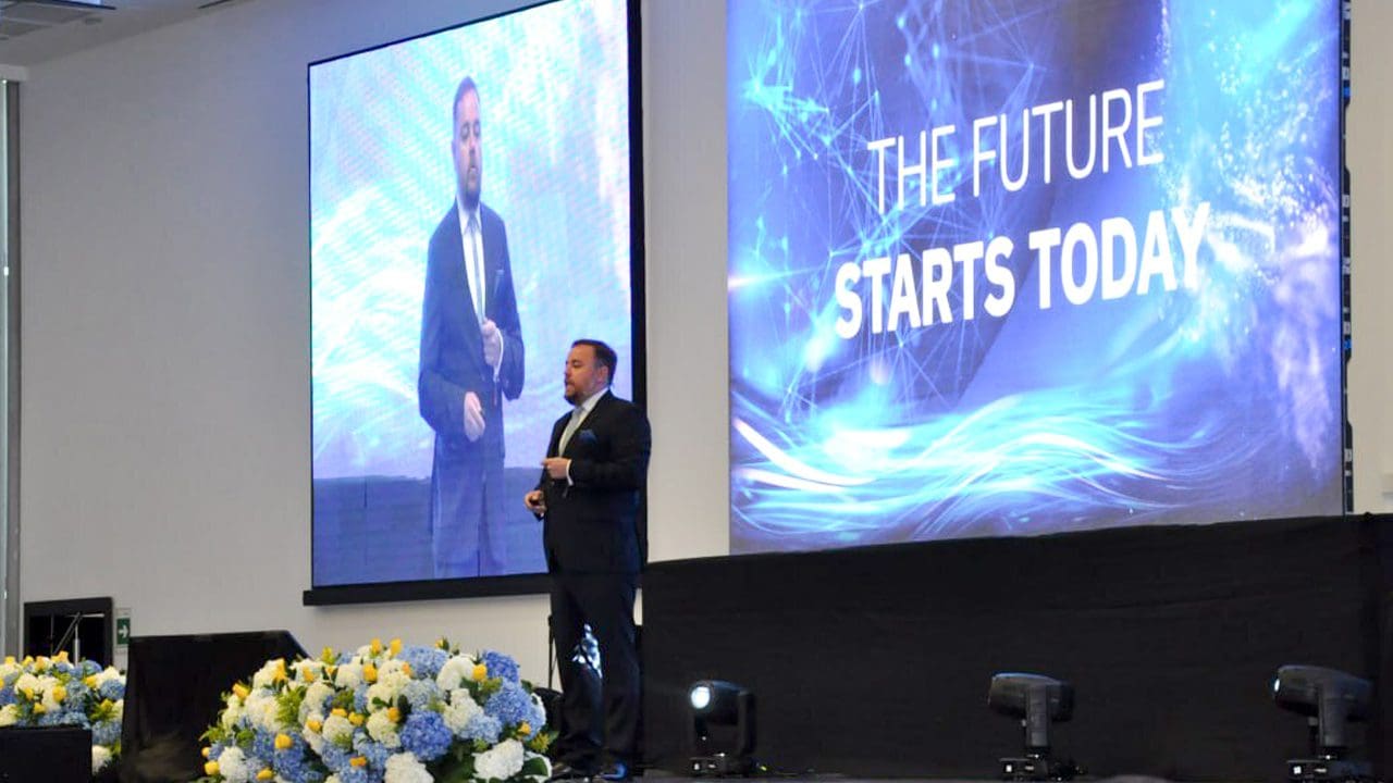 Um homem de terno está no palco ao lado de um telão. As palavras "O FUTURO COMEÇA HOJE" são projetadas na tela. Ele parece estar fazendo uma apresentação em um evento corporativo da OES. O palco está decorado com flores.