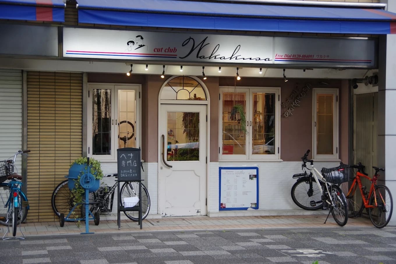 A loja tem uma placa que diz "cut club Wakana" em cima de uma porta dupla branca, e oferece serviços de atalho e henna. Entalados entre as janelas e o estacionamento de bicicletas, há bicicletários azuis, plantas de casa e menus e placas perto da entrada.