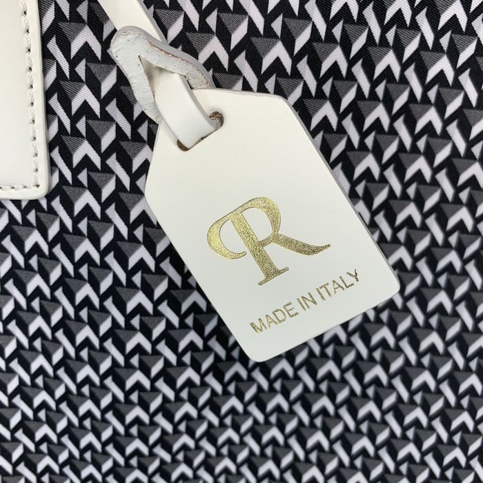 Uma etiqueta de couro branco ROBERTA PIERI com as letras "R" em ouro e as palavras "MADE IN ITALY" está pendurada no fundo de um tecido geométrico preto e branco.