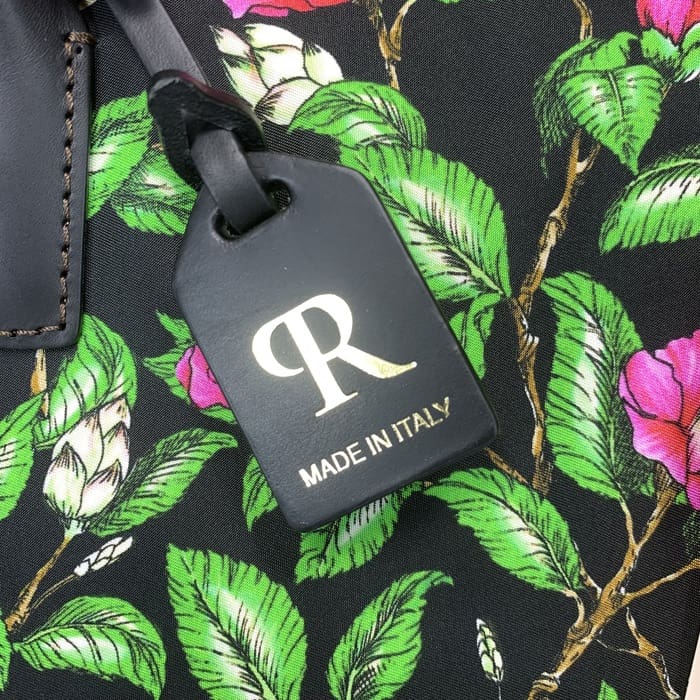 A etiqueta de bagagem preta ROBERTA PIERI com o "R" dourado e texto italiano é suspensa a partir de um tecido floral com folhas verdes e flores rosas, complementando o mini duffel com elegância italiana intemporal.