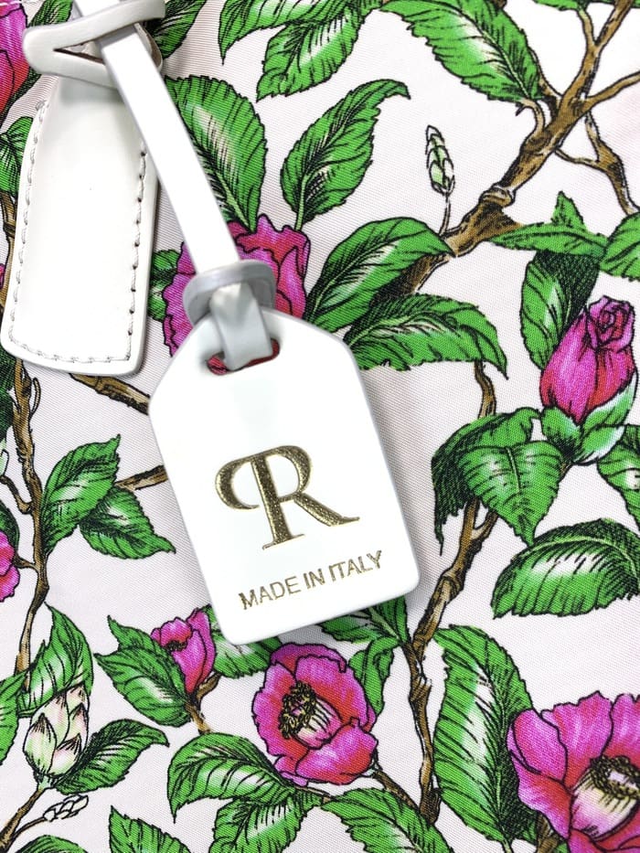 Close-up da mini bolsa duffel ROBERTA PIERI. pedúnculos com folhas verdes e flores rosas; A etiqueta branca anexada ao saco de 2 vias apresenta um logotipo "R" dourado e as palavras "MADE IN ITALY".